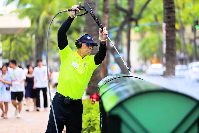 A man in a neon uniform pressure washes some newspaper receptacles. 