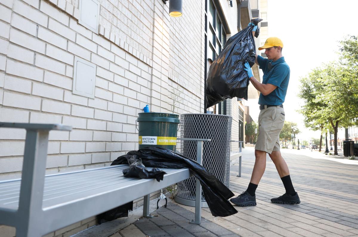 An Arlington Ambassador pulls a trash bag out from a metal trash can along a brick wall on a sidewalk.