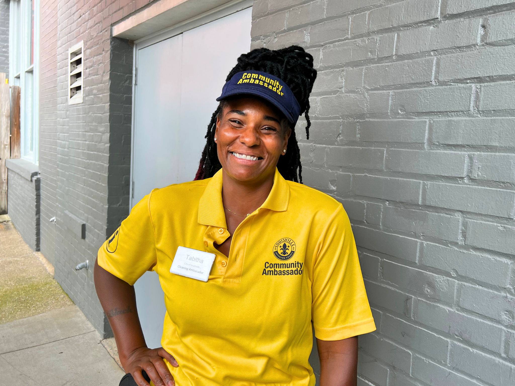 A woman in a yellow uniform smiles for the camera against a grey brick wall.